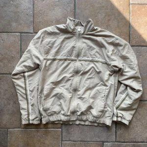 Beige vindjacka från Nike - Säljer en beige vindjacka från Nike i storlek S. Jackan har en dragkedja framtill och är långärmad med elastiska muddar. Perfekt för vår och höst med sin lätta och bekväma passform. Nike-loggan är broderad på bröstet och dragkedjan har en snygg Nike-