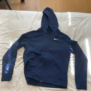 Säljer en mörkblå hoodie från Nike i storlek barn XL. Den är gjord av mjukt och varmt Therma-FIT-material, perfekt för kyliga dagar. Hoodien har en dragkedja framtill och en klassisk Nike-logga på bröstet. Den är i bra skick och redo för nya äventyr!