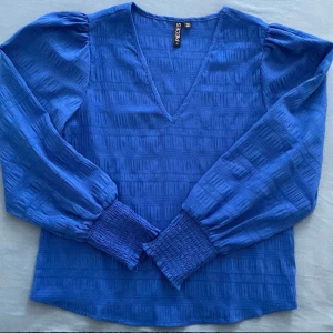 Blus från Pieces - En blå blus med långa ärmar och puffärmar. Blusen har en V-ringning och är gjord av ett mjukt, lätt tyg. Den har en elastisk detalj vid ärmsluten. I bra skick.