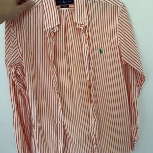 Randig skjorta från Ralph Lauren - Säljer en snygg och stilren randig skjorta från Ralph Lauren. Den är vit med orange ränder och har en klassisk krage. Skjortan är slim fit och har långa ärmar med knappar. Perfekt för både vardag och fest!