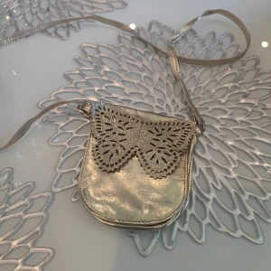 Fjärilsformad axelremsväska i guld - Söt liten axelremsväska i guld med fjärilsdetalj. Perfekt för att förvara småsaker som mobiltelefon, plånbok och nycklar.