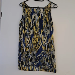 Mönstrad ärmlös klänning - Säljer en superfin ärmlös troja med ett coolt mönster i blått, gult och svart. Perfekt för sommardagar eller en festlig kväll. Den har en lös passform och är gjord i ett lätt och bekvämt material.