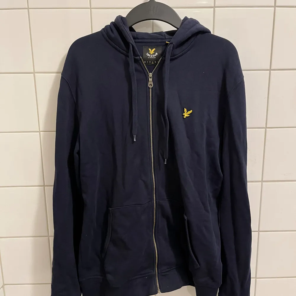 Säljer en snygg och bekväm mörkblå hoodie från Lyle & Scott. Den har en dragkedja framtill och en broderad gul logga på bröstet. Perfekt för en avslappnad stil och passar till alla tillfällen. Den köptes för 1200 kr och har använts med försiktighet. Hör av dig vid intresse!. Hoodies.