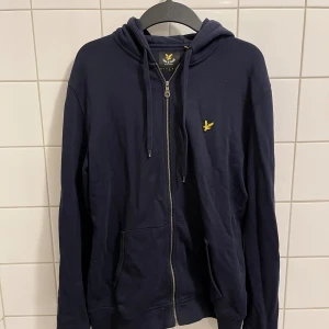 Mörkblå hoodie från Lyle & Scott - Säljer en snygg och bekväm mörkblå hoodie från Lyle & Scott. Den har en dragkedja framtill och en broderad gul logga på bröstet. Perfekt för en avslappnad stil och passar till alla tillfällen. Den köptes för 1200 kr och har använts med försiktighet. Hör av dig vid intresse!