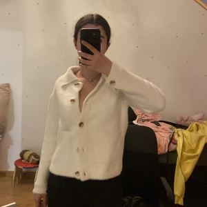 Vit kofta med knappar - Säljer en supermysig vit kofta med stora knappar framtill. Den har en klassisk krage och långa ärmar, perfekt för kyliga dagar. Koftan är i ett mjukt material som känns skönt mot huden. Den har också praktiska fickor fram. Perfekt för både vardag och lite finare tillfällen!