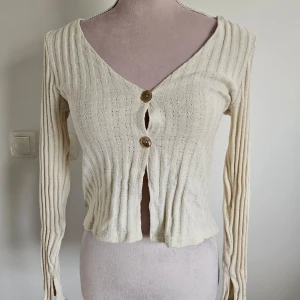 Beige ribbad kofta från NLY Trend - Säljer en superfin beige kofta från NLY Trend i storlek M. Den är ribbad och har en v-ringad design med två snygga knappar framtill. Perfekt för höst och vår, och den är långärmad vilket gör den mysig och stilren. 