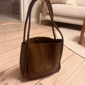 Brun axelväska i skinn - Säljer en snygg brun axelväska i skinn. Den har en stilren design med långa remmar och en praktisk storlek som passar perfekt för vardagsbruk. Väskan har en enkel knappstängning upptill och är i mycket fint skick.