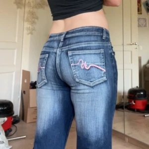 Blå bootcut jeans med rosa broderi - Säljer ett par snygga blå bootcut jeans med coolt rosa broderi på bakfickorna. De är lågmidjade och har en skön passform som sitter perfekt!  Mått: Innerbenslängden: ca 80  Midjemåttet: ca 42.5  ☺️⭐️