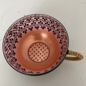 Vacker kopp med mönster - Säljer en superfin kopp med ett detaljerat mönster i kopparfärgad och rosa. Insidan har ett prickigt mönster och utsidan är svart. Handtaget är flätat och ger en rustik känsla.Mockakoppar i emalj från Österrike.höjd 7cm . djupt 3 cm