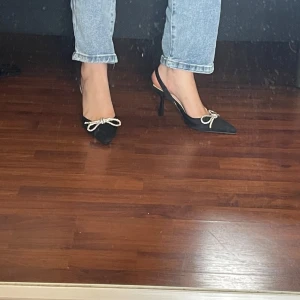 Klackskor  - Säljer ett par eleganta svarta pumps med spetsig tå och glittriga rosetter framtill. Perfekta för festliga tillfällen! De har en stilren design med en smal rem runt hälen för extra stöd. Storlek 39. Enbart testade