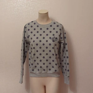 Grå prickig tröja med mönsterdetalj - Säljer en superfin grå tröja med mörkblå prickar över hela. Den har en unik mönsterdetalj på bröstet som ger den lite extra stil. Tröjan är långärmad och perfekt för både vardag och chill. Den är i bra skick och redo för en ny ägare!