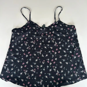 Svart blommig linnetopp - Säljer en superfin svart linnetopp med små blommor i olika färger. Den är perfekt för sommardagar och har smala axelband. Materialet känns lätt och luftigt, vilket gör den idealisk för varma dagar. Passar både till vardags och fest!