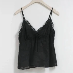 Svart spetslinne med volangdetaljer - Säljer ett snyggt svart linne med tunna axelband och v-ringning. Linnet har fina spetsdetaljer och en liten rosett framtill. Perfekt för en festlig kväll eller en somrig dag.