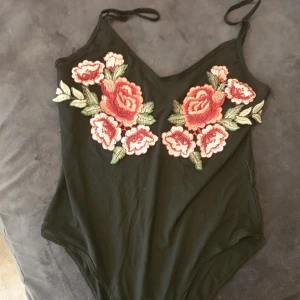 Svart bodysuit med blommigt broderi - Säljer en snygg svart body med tunna axelband och v-ringad design. Den har ett vackert blommigt broderi i rosa och grönt framtill. Perfekt för en festlig kväll eller en utekväll med vänner! den är genomklinlig och storlek s.