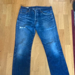 Levi's Premium jeans - Säljer ett par snygga Levi's jeans i blå denim. De har en klassisk straight fit och är i välanvänt skick med en cool slitning på ena benet. Perfekta för en avslappnad stil. Knapparna framtill ger en vintagekänsla.