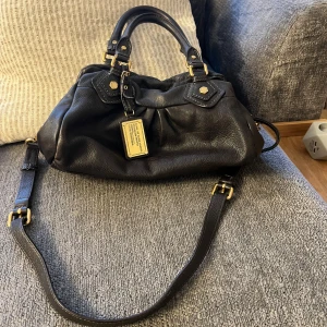 Marc Jacobs väska  - Mått : 30x19 cm dustbag ingår 🩷Snygg mörk brunt axelväska i skinn med gulddetaljer och justerbar axelrem. Väskan har en dragkedja och ett mönstrat innerfoder med logotyp. Perfekt för att bära med sig det viktigaste med stil. Fraktas eller möter upp i centrala Stockholm 🩷 