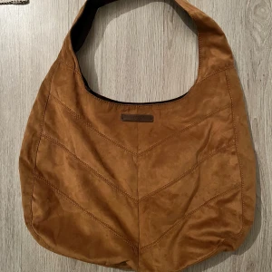 Brun axelväska från Esprit - Snygg brun axelväska från Esprit i mocka imitation. Väskan har ett litet fack med dragkedja och en bekväm axelrem. Perfekt för att bära dina dagliga nödvändigheter med stil.