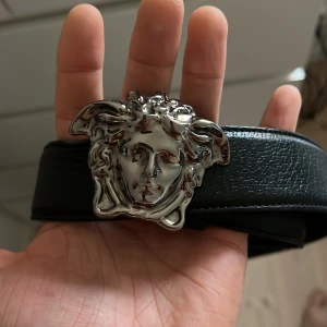 Svart skärp med silverdetalj från Versace - Snyggt svart skärp i skinn från Versace med en iögonfallande silverspänne i form av ett huvud. Perfekt för att ge en lyxig touch till din outfit.
