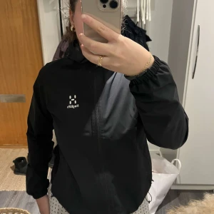 Svart vindjacka från Haglöfs - Snygg svart vindjacka från Haglöfs med dragkedja och huva. Perfekt för blåsiga dagar och utomhusaktiviteter. Jackan har en stilren design med Haglöfs-logga på bröstet. säljer pga att den inte kommer till användning, mycket bra skick och passar både S och M