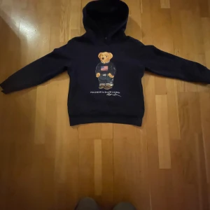 Mörkblå hoodie från Ralph Lauren - Säljer en snygg mörkblå hoodie från Ralph Lauren med en björn på framsidan. Hoodien har långa ärmar och en klassisk huva. Perfekt för en avslappnad stil.