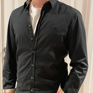 Svart skjorta från Burberry Brit - Snygg svart skjorta från Burberry Brit med klassiska knappar och stilren design. Perfekt för en elegant look. Den har långa ärmar och en diskret logga på bröstet.