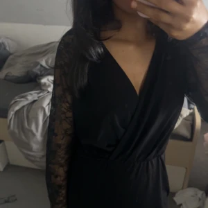 Svart klänning med spetsärmar från Vero Moda - Elegant svart klänning från Vero Moda med v-ringad omlott-design och spetsdetaljer på ärmarna. Perfekt för en stilren look.