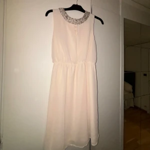 Vit ärmlös klänning med paljettkrage - Elegant vit klänning med en glittrig paljettkrage. Den är ärmlös och har en kort längd, perfekt för festliga tillfällen. Klänningen har en lätt och luftig passform med en subtil öppning framtill vid halsen.