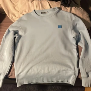 Ljusblå tröja från Acne Studios - Säljer en stilren ljusblå tröja från Acne Studios i 100% bomull. Tröjan har en enkel design med en liten fyrkantig logga på bröstet och långa ärmar. Perfekt för en avslappnad look.