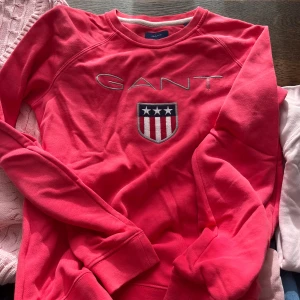 Hallonröd  sweatshirt från Gant - Snygg Hallonröd sweatshirt från Gant med broderad logga och sköld på bröstet. Tröjan har långa ärmar och rund halsringning. Perfekt för en avslappnad stil.