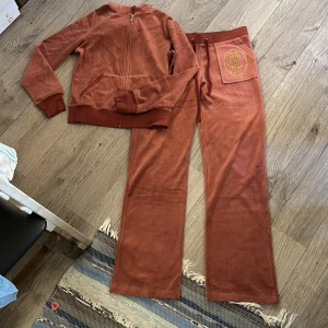 Rostfärgad velourtränings set från Juicy Couture - Säljer en snygg och bekväm rostfärgad velourtränings set från Juicy Couture. Setet består av en hoodie med dragkedja och matchande byxor med resår och snörning i midjan. Båda delarna har Juicy Coutures logga i guld på baksidan och byxan. Perfekt för en avslappnad stil. Uppskattad storlek M inköpt nyligen på tradera. 