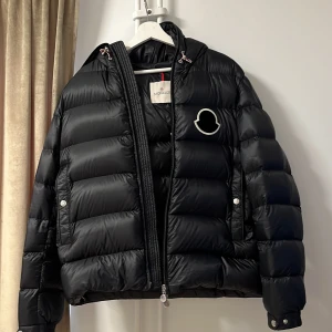 MONCLER SASSIERE  - Snygg svart dunjacka från Moncler med en stilren design. Jackan har en dragkedja framtill och en huva för extra värme. Den är quiltad och har ett broderat märke på bröstet. Perfekt för kyliga dagar.
