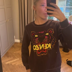 Svart lila sweatshirt från Kenzo med tigerbroderi - Säljer en svart sweatshirt från Kenzo med ett färgglatt tigerbroderi på framsidan. Tröjan har långa ärmar och en rund halsringning. Perfekt för en cool och avslappnad stil.