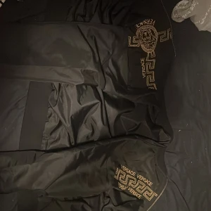 Svart jacka från Versace - Snygg svart jacka från Versace med guldiga detaljer och logotyp. Jackan har dragkedja och är tillverkad i 100% polyester. Perfekt för en stilren look.