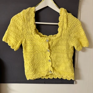 Gul stickad topp från Zara - Säljer en söt gul stickad topp från Zara med korta ärmar och dekorativa knappar framtill. Toppen har en vågig kant och en fin struktur i materialet. Perfekt för en somrig look! 🌼