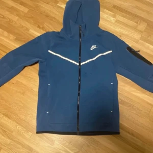 Blå hoodie från Nike - Säljer en blå hoodie från Nike med dragkedja och huva. Den har vita detaljer och en ficka på ärmen. Koftan har fått några småfläckar på sig, har dock inte testat att få bort💕💕 men dom syns knappast