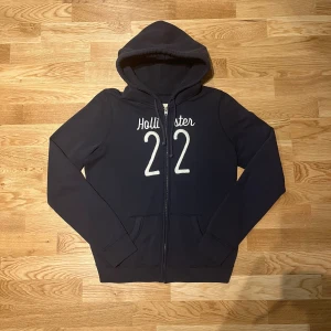 Mörkblå hoodie från Hollister - Säljer en mörkblå dam hoodie från Hollister med dragkedja och tryck 'Hollister 22' på framsidan. Tröjan har en bekväm huva och långa ärmar, perfekt för en avslappnad stil.
