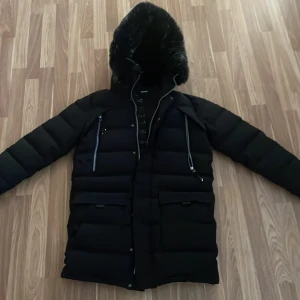 Svart dunjacka från Supply & Demand - Snygg svart dunjacka från Supply & Demand med pälskantad huva och dragkedja. Jackan har flera praktiska fickor och en stilren design som passar perfekt för kalla dagar.
