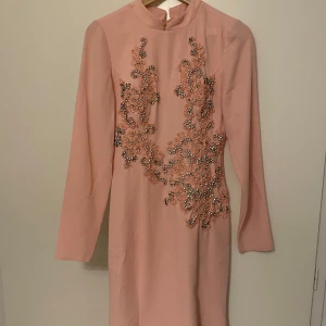 Rosa långklänning med broderade detaljer - Elegant rosa långklänning med långa ärmar och vackra broderade detaljer i form av blommor och pärlor. Klänningen har en hög hals och en figursydd passform som ger en stilren siluett. Passar strl 36-38 (S/M). Nypris 2000kr