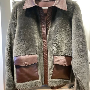 Shearling jacka från Remain - Snygg grå pälsjacka från Remain med bruna lamm läderdetaljer. Jackan har en klassisk krage och stora fickor med knappar framtill. Perfekt för kyliga dagar när du vill vara både varm och stilren.