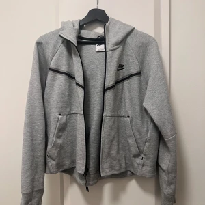 Grå hoodie från Nike - Säljer en  grå tech hoodie från Nike med dragkedja och svarta detaljer. Tröjan har en klassisk passform med långa ärmar och praktiska fickor framtill.