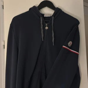 Mörkblå hoodie från Moncler - Säljer en snygg mörkblå hoodie från Moncler med dragkedja och huva. Tröjan har en broderad Moncler-logga på ärmen och detaljer i rött och vitt. Perfekt för en stilren look. Tröjan är i storlek xxl men sitter som en large, tröjan är i gott skick men använd. 