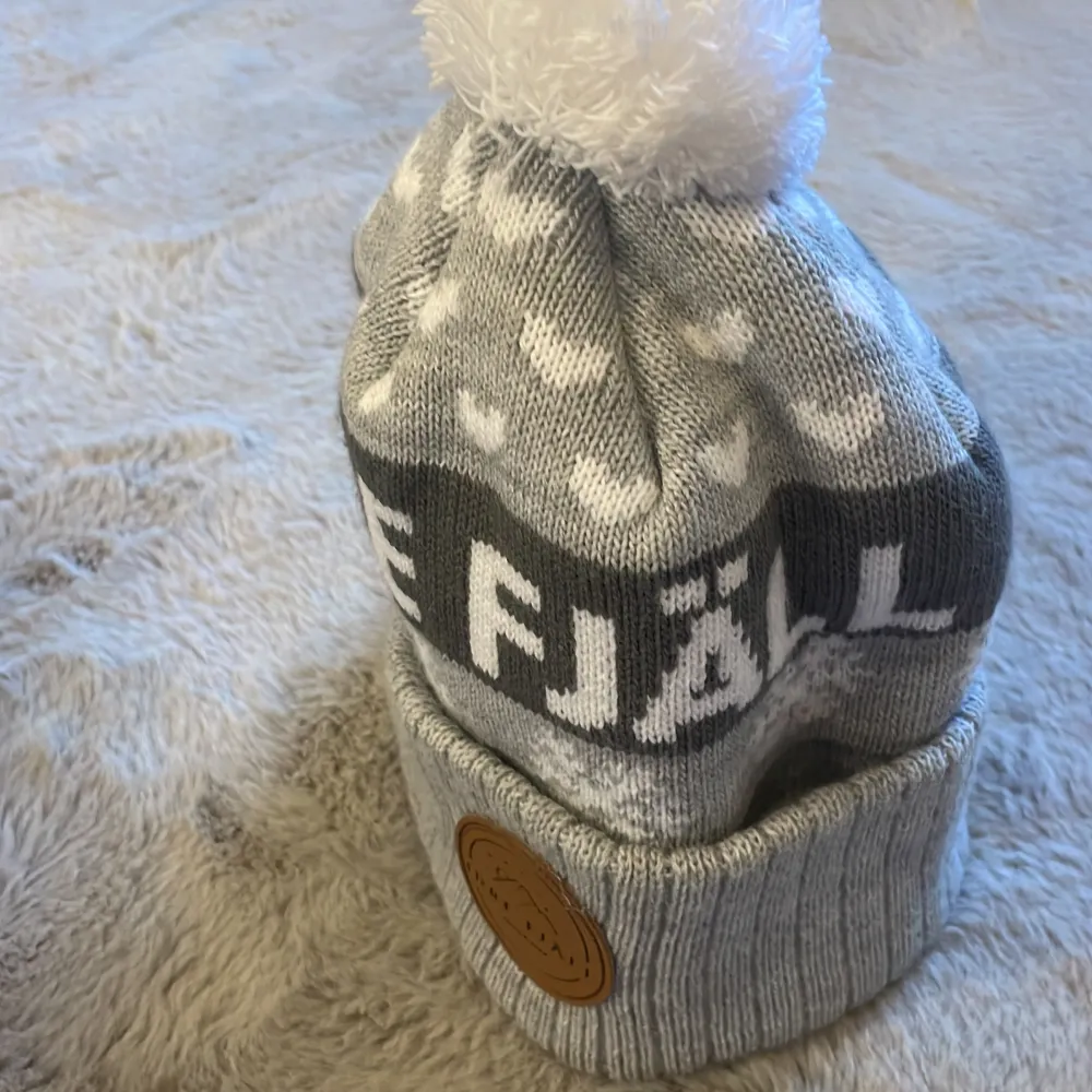 Snygg grå stickad mössa från Idre Fjäll med vit tofs och snöflingemönster. Mössan har en uppvikt kant med ett brunt märke i skinnimitation. Perfekt för kyliga dagar! Köpt i Idre vintern 2025. Asusteet.