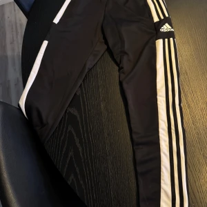 Svarta träningsbyxor från Adidas - Snygga svarta träningsbyxor från Adidas med de klassiska tre vita ränderna längs sidorna. Byxorna har en elastisk midja och är perfekta för träning eller en avslappnad stil. Adidas-loggan är tryckt på framsidan.
