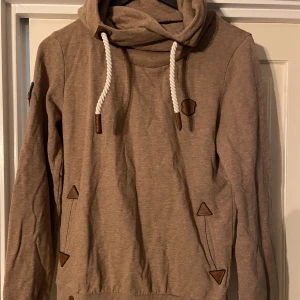 Beige hoodie från Naketano - Säljer en beige hoodie från Naketano med en mysig krage och vita dragsnören. Tröjan har långa ärmar och dekorativa bruna detaljer vid fickorna och kragen. Perfekt för en avslappnad stil.