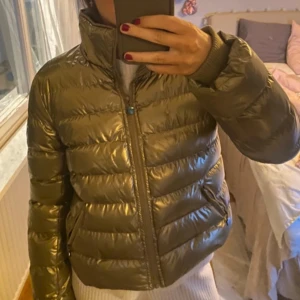 Guld pufferjacka med emblem - Snygg guld pufferjacka med hög krage och dragkedja. Jackan har ett coolt emblem på ärmen och ger en glansig look. Perfekt för att hålla värmen med stil.