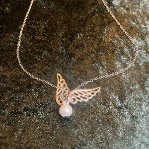 Halsband med vingar och pärla - Elegant halsband med en fin design av vingar och en vit pärla. Kedjan är tunn och smidig, vilket ger ett stilrent intryck. Perfekt för att lägga till en touch av elegans till din outfit.