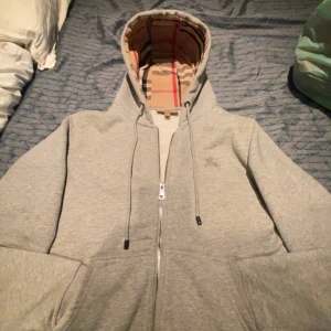 Burberry hoodie - äkta och i toppskick! - Säljer min grå Burberry hoodie med den klassiska rutiga fodringen i luvan. Storlek S, unisex. Hoodien är i mycket fint skick utan några fläckar eller skador.  Detaljer: 	•	Äkta Burberry (se bilder för detaljer på dragkedja och logotyp) 	•	Mjuk bomull, perfekt för både vardag och lite finare tillfällen 	•	Nypris: ca 6 000 kr 	•	Mitt pris: 1 299 kr (kan diskuteras vid snabb affär)