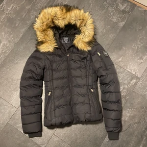 Svart dunjacka från Vinson Polo Club - Säljer denna snygga svarta dunjacka från Vinson Polo Club med pälskantad huva och dragkedja. Den är superskön och håller verkligen värmen. Använd endast en vinter och är i väldigt bra skick.