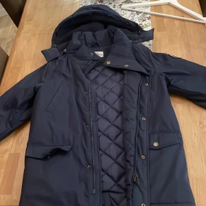 Mörkblå jacka från Hampton Republic - Snygg mörkblå jacka från Hampton Republic med quiltad insida och avtagbar huva. Jackan har praktiska fickor med knappar och stängs med dragkedja och knappar framtill. Perfekt för kyligare dagar.