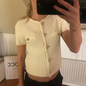 Vit stickad kofta med guldknappar - Säljer en söt vit stickad kofta från H&M med korta ärmar och dekorativa guldknappar. Koftan har två små fickor framtill och en ribbad kant vid ärmslut och nederkant. Perfekt för en stilren look! Köpt för 400 kr och använd få tal gånger 💞